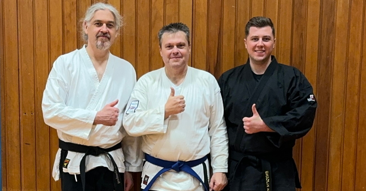 Drei Shaolin-Kempoka, zwei auf der linken Seite mit weißer Jacke und einer mit schwarzer Jacke rechts. Alle zeigen Daumen-hoch.