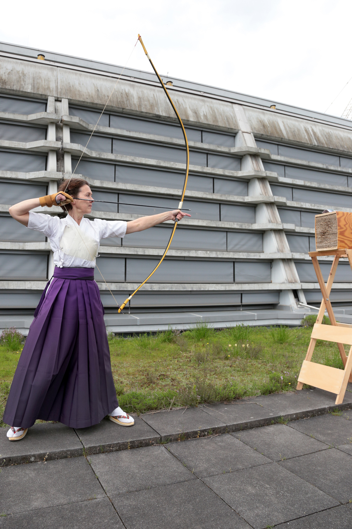 Bild mit Monika Nadler mit gespanntem Bogen vor einem Makiwara. Im Hintergrund eine Betonwand.